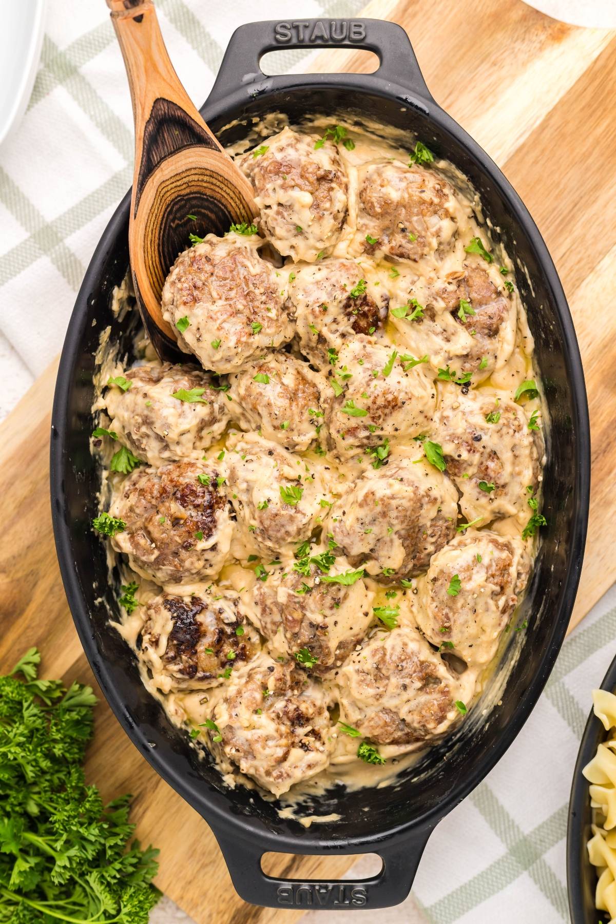 Gluten free Swedish meatballs in creamy sauce, garnished with parsley, served in a black oval dish with a wooden spoon.