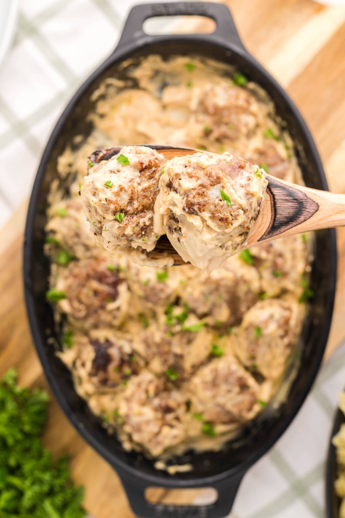 Wooden spoon holding creamy gluten free Swedish meatballs over a black dish filled with more meatballs in sauce, garnished with herbs.