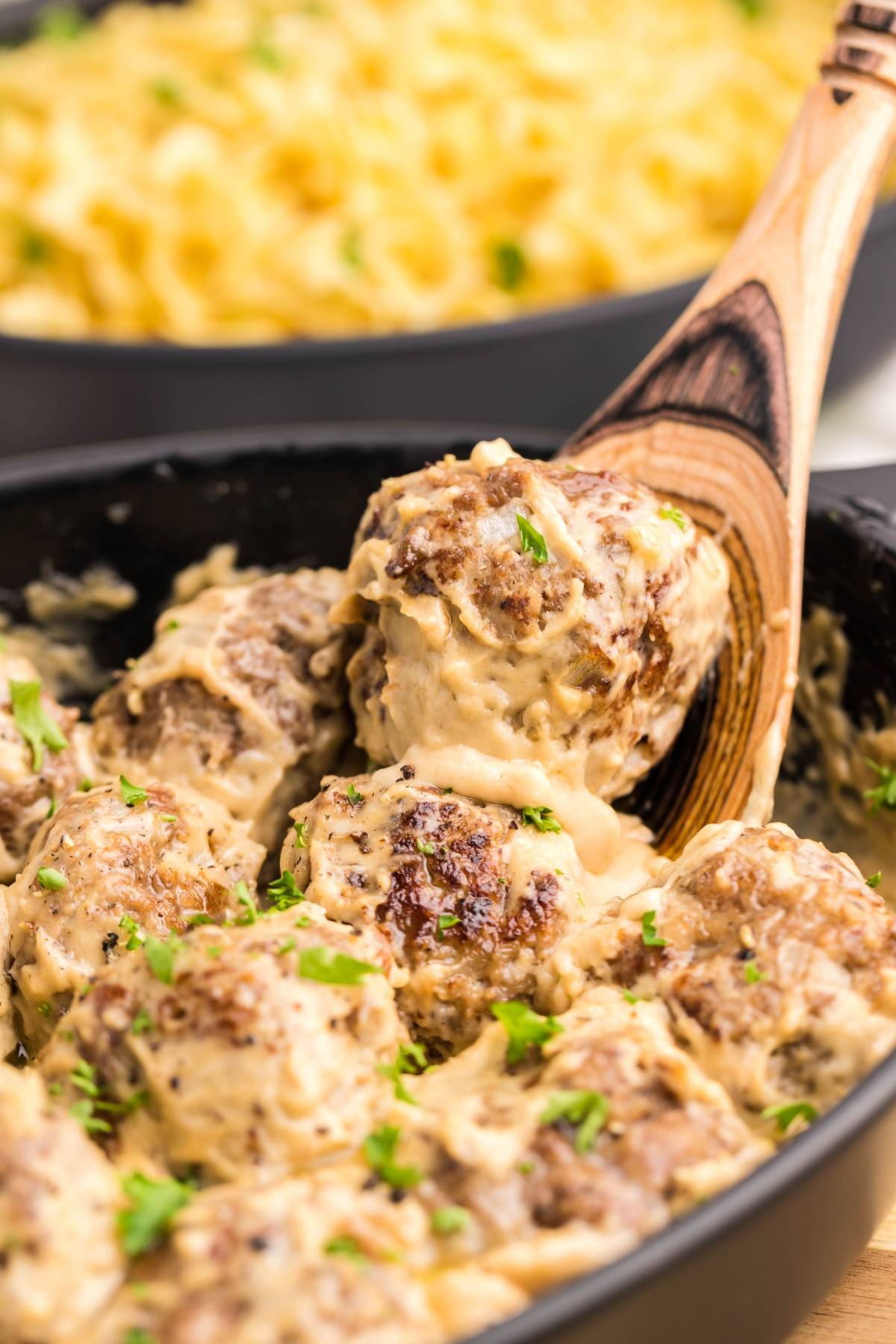 Meatballs in creamy sauce being served with a wooden spoon, garnished with parsley. Mac and cheese in background—just like a Copycat Ikea Swedish meatballs recipe you can make at home.