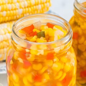 Two jars filled with homemade corn relish are surrounded by fresh corn on the cob and diced vegetables.