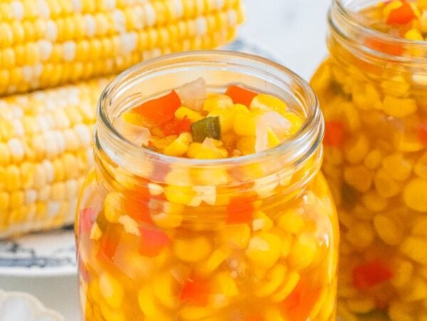 Two jars filled with homemade corn relish are surrounded by fresh corn on the cob and diced vegetables.