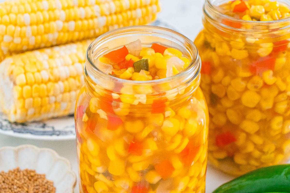 Two jars of homemade corn relish with chopped red peppers and onions, alongside fresh corn cobs in the background.