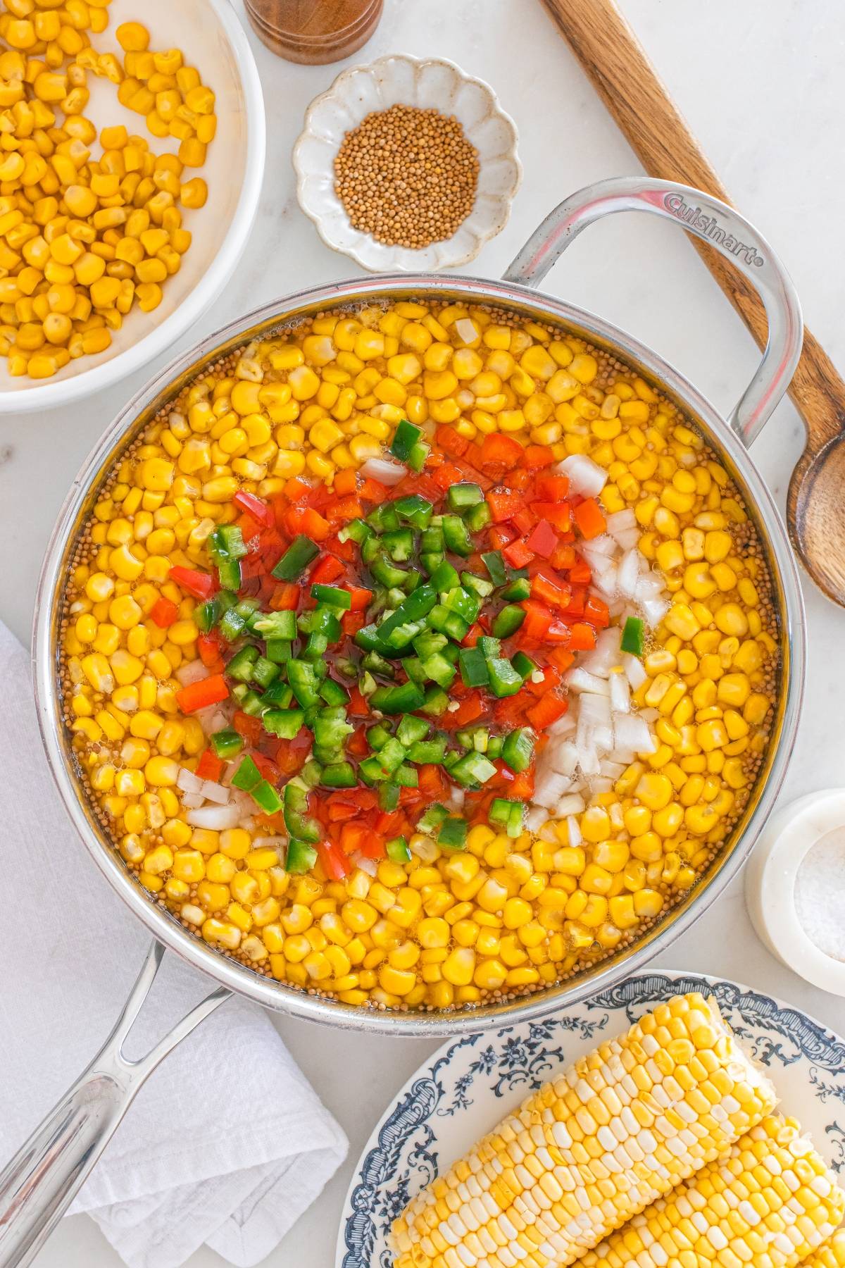 A skillet filled with corn, diced red bell pepper, jalapeño, and onion, surrounded by ingredients on a counter.