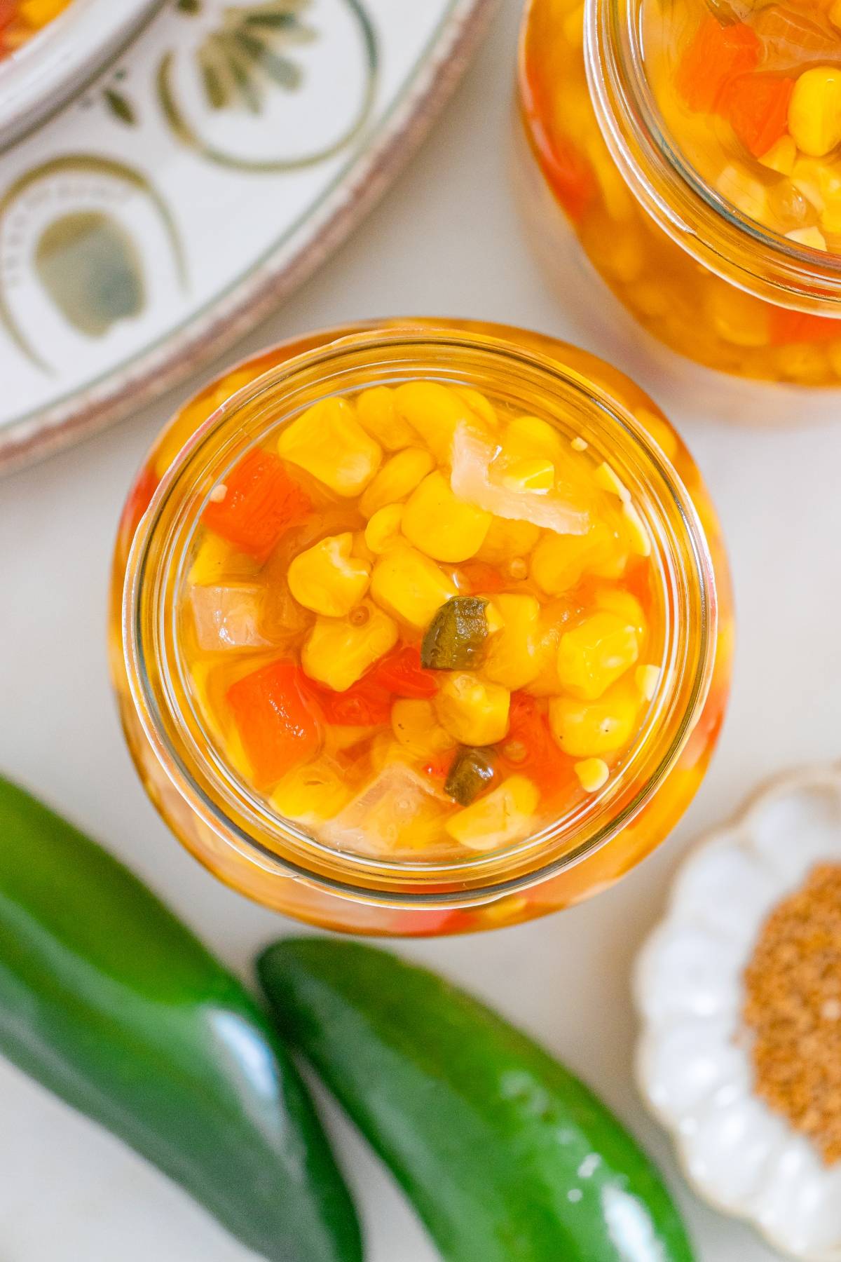 Overhead view of a jar filled with corn relish, surrounded by green peppers and a dish of spices.