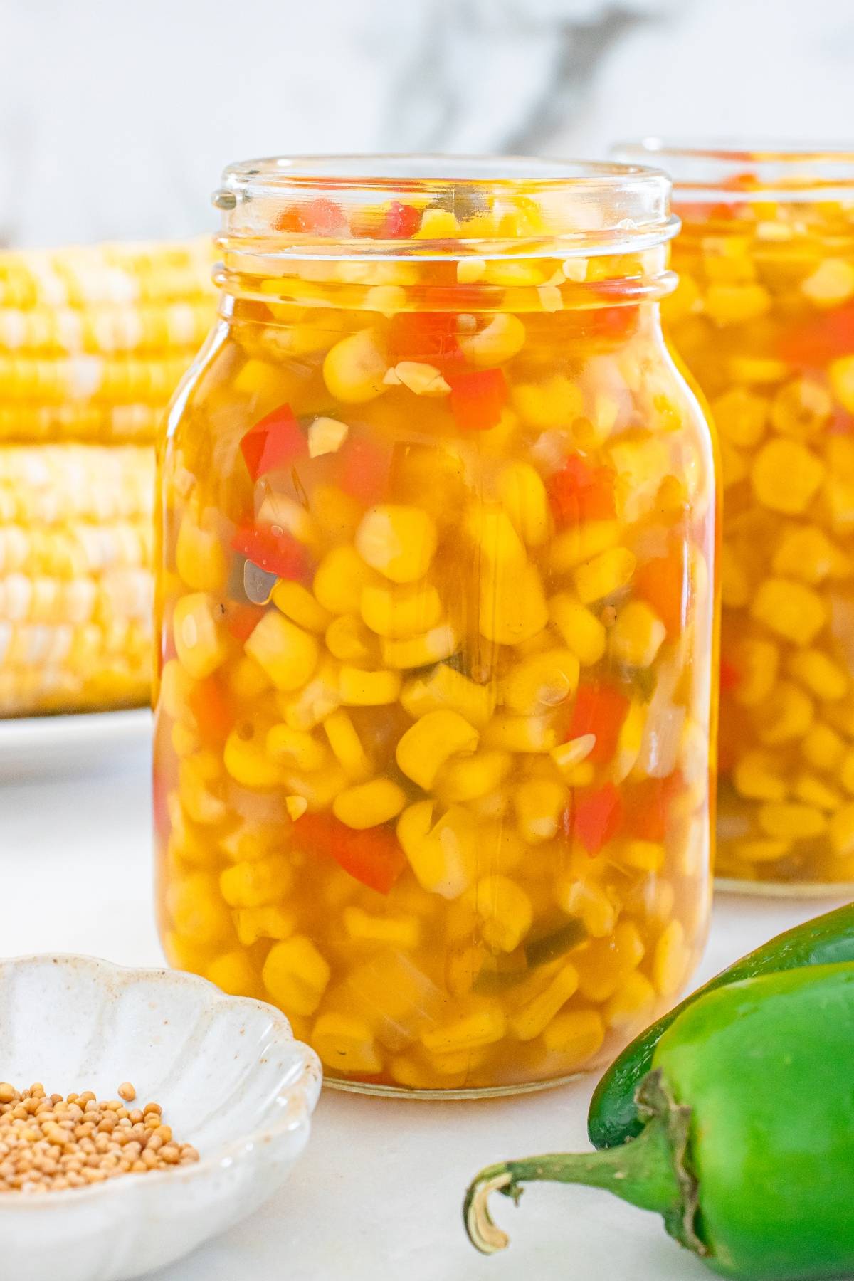 A jar filled with pickled corn and diced red peppers, with corn cobs and a jalapeño in the background.
