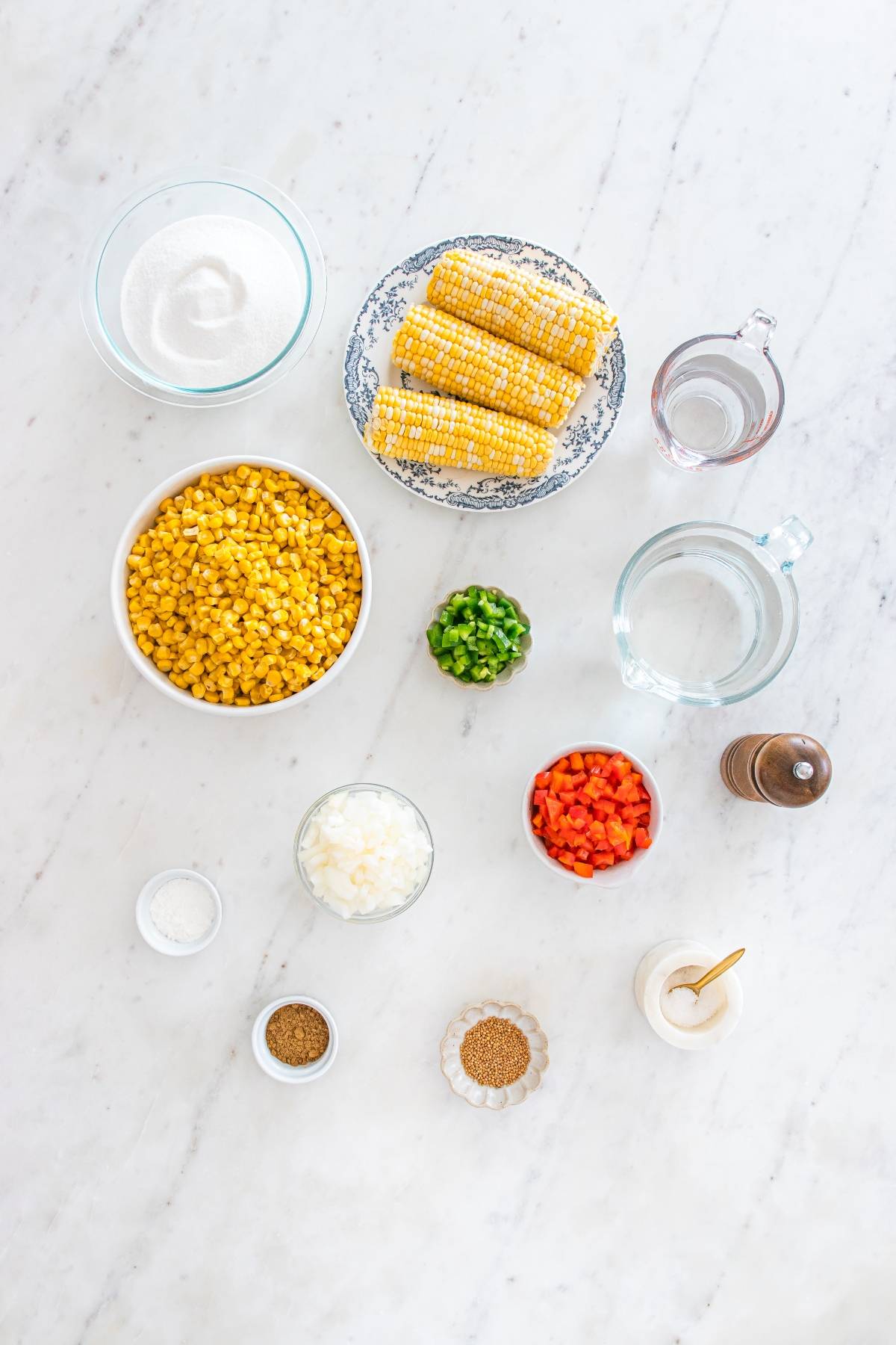 Ingredients for corn soup arranged on a white surface, including corn, peppers, onion, spices, and water.