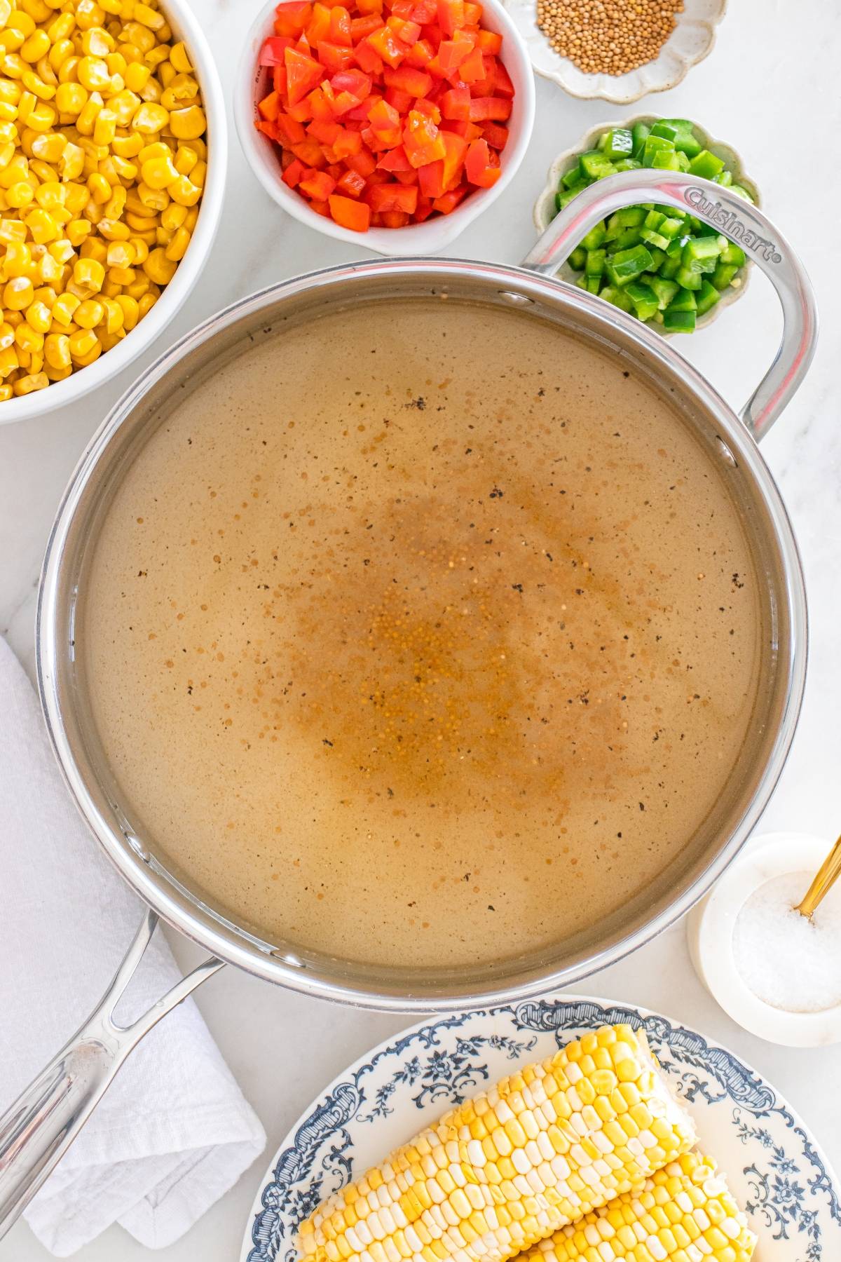 A pot of seasoned broth surrounded by corn, chopped tomatoes, green peppers, and corn cobs on a counter.