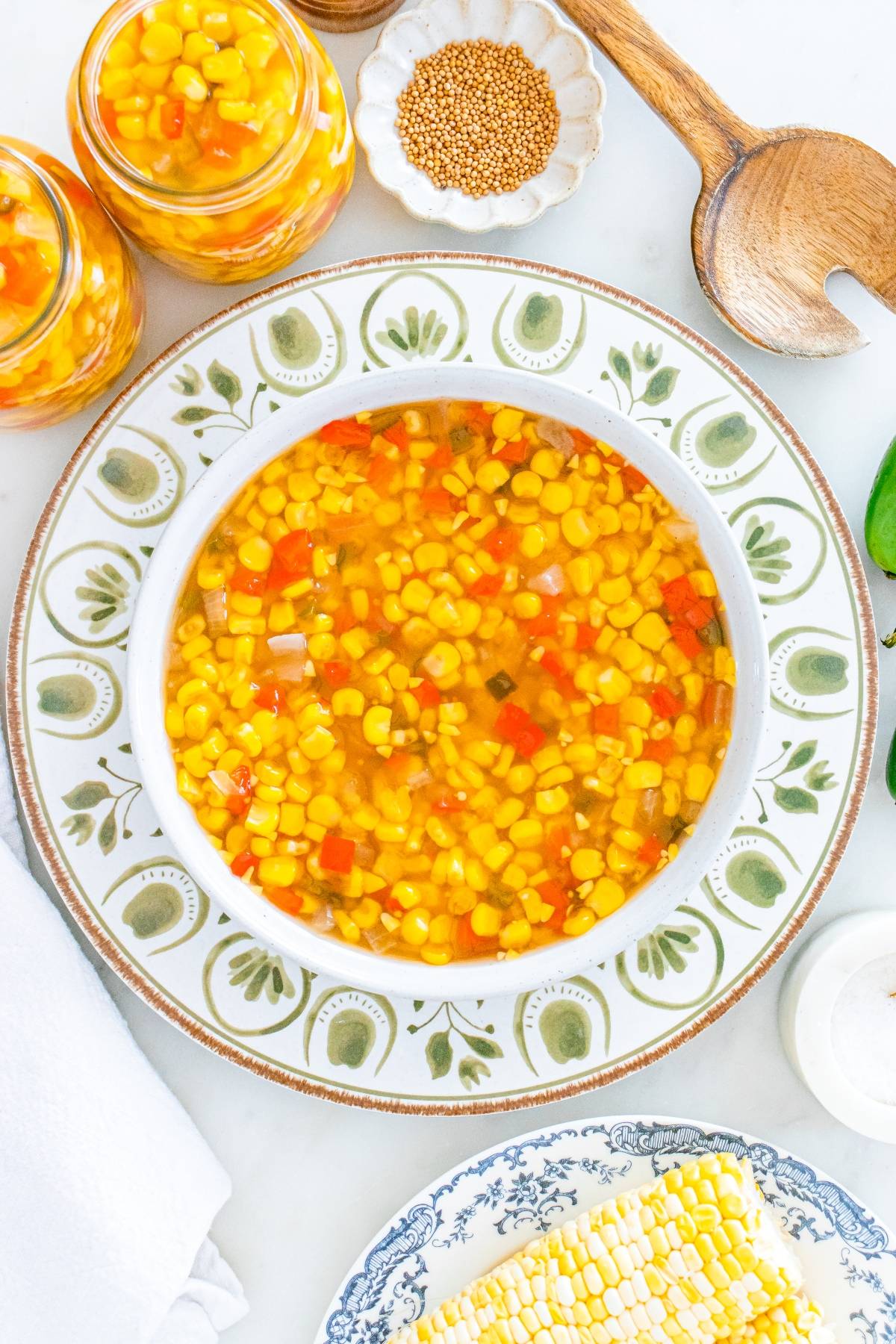A bowl of corn relish with chopped vegetables, surrounded by corn, jars, and wooden utensils on a table.