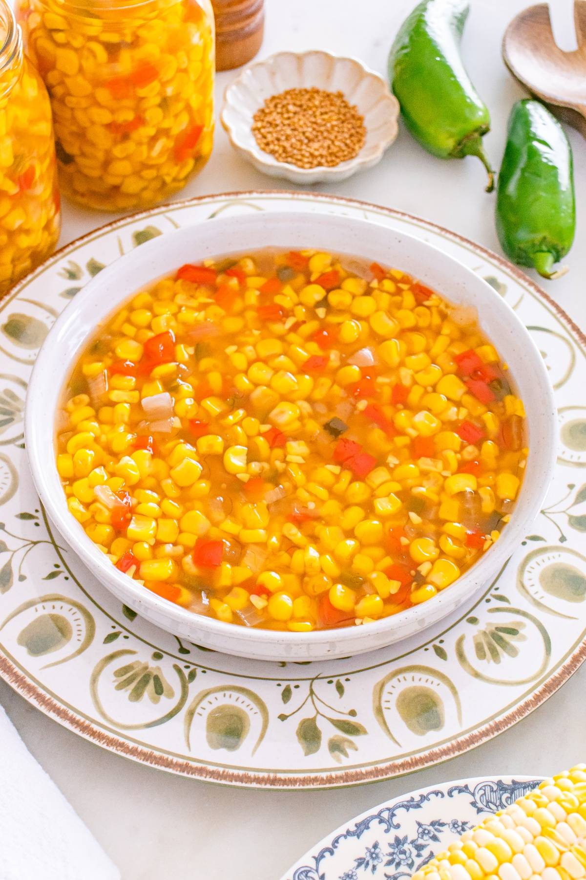 A bowl of corn relish with red peppers and onions sits on a decorative plate, with jars and jalapeños nearby.