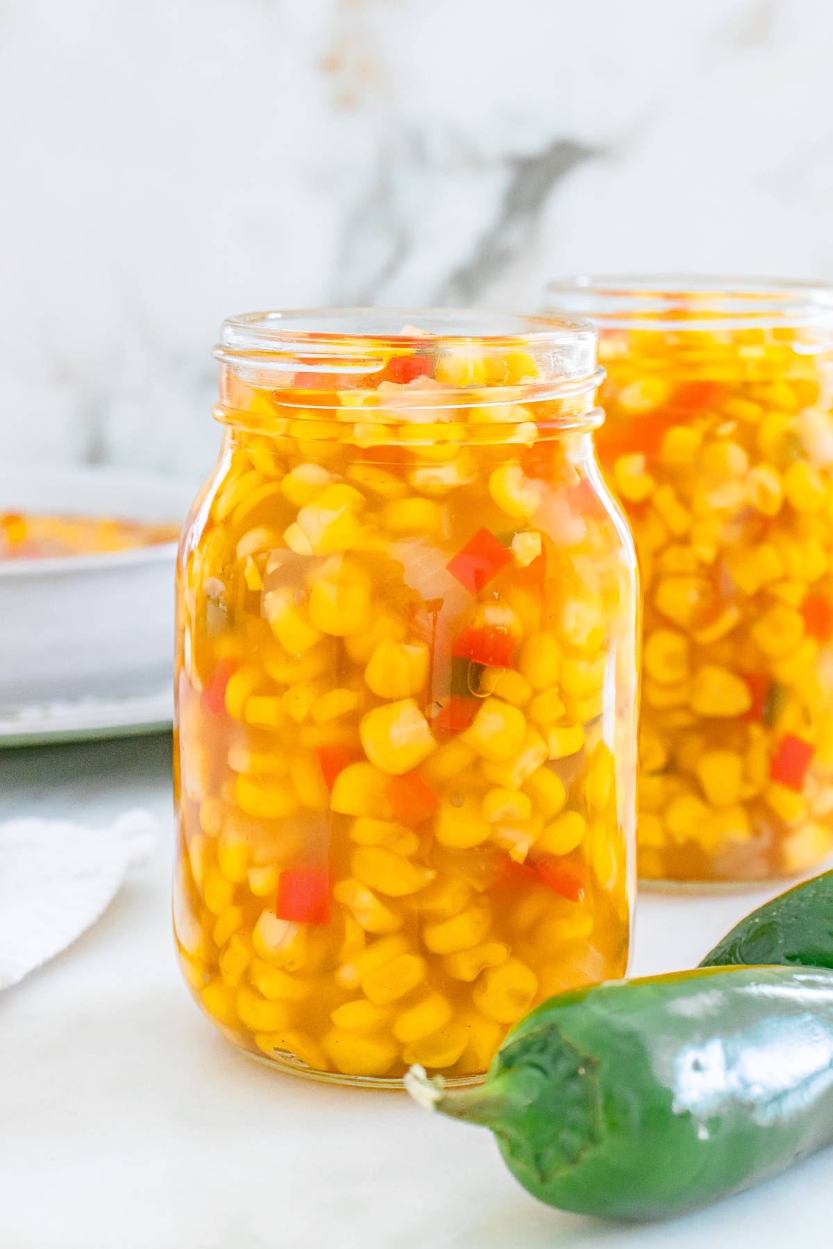 A glass jar filled with homemade corn relish, with diced red peppers, next to a jalapeño pepper.
