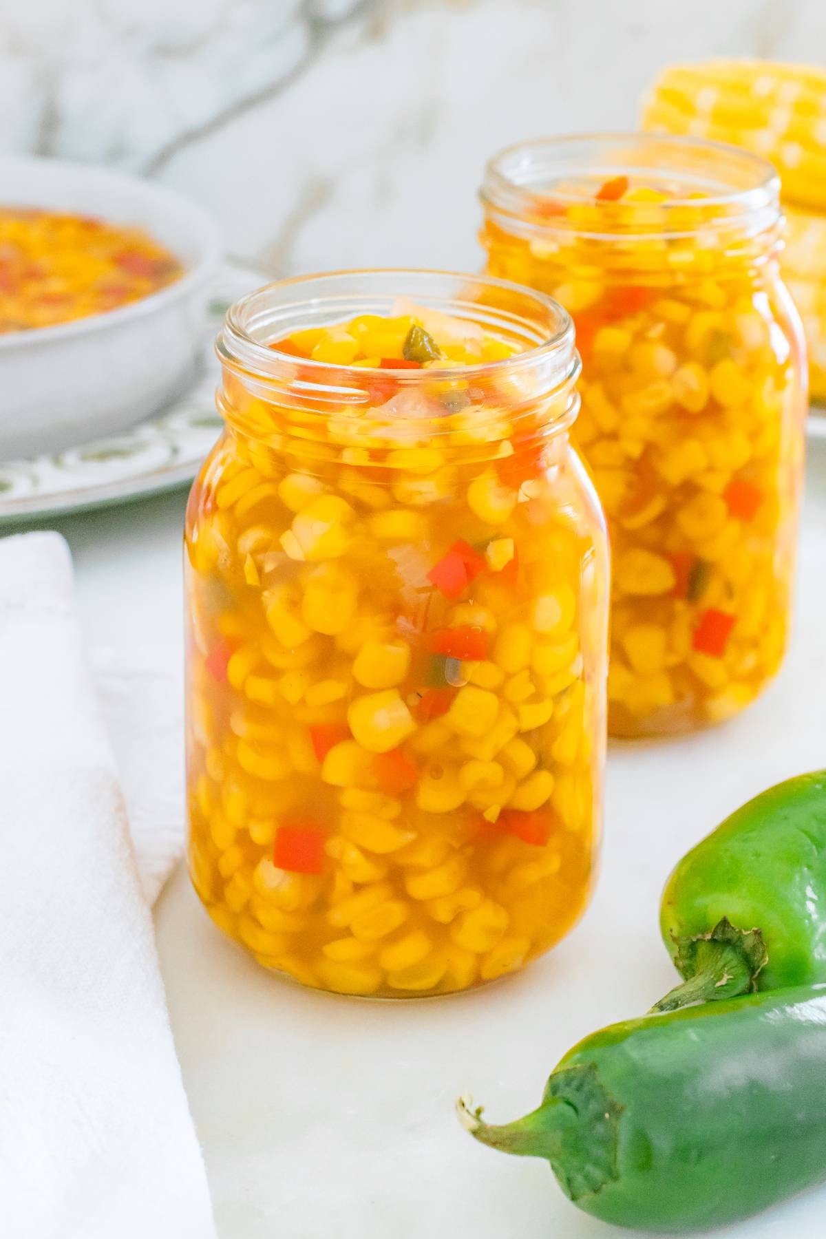 Two jars filled with pickled corn and diced red peppers, with fresh jalapeños and corn in the background.