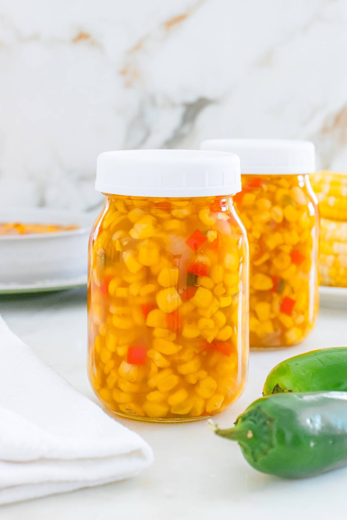 Two jars filled with corn relish, with diced red peppers, on a white surface next to fresh green jalapeños.