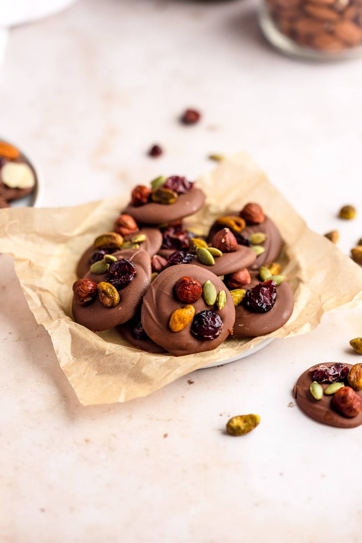 Chocolate disks topped with dried fruit and nuts, reminiscent of cranberry nut clusters, arranged on parchment paper on a light surface.