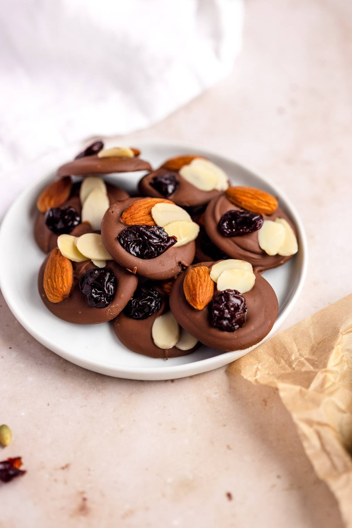 A plate of chocolate disks topped with almonds, dried cherries, and sliced almonds on a light surface, perfect for fans of cranberry nut clusters.