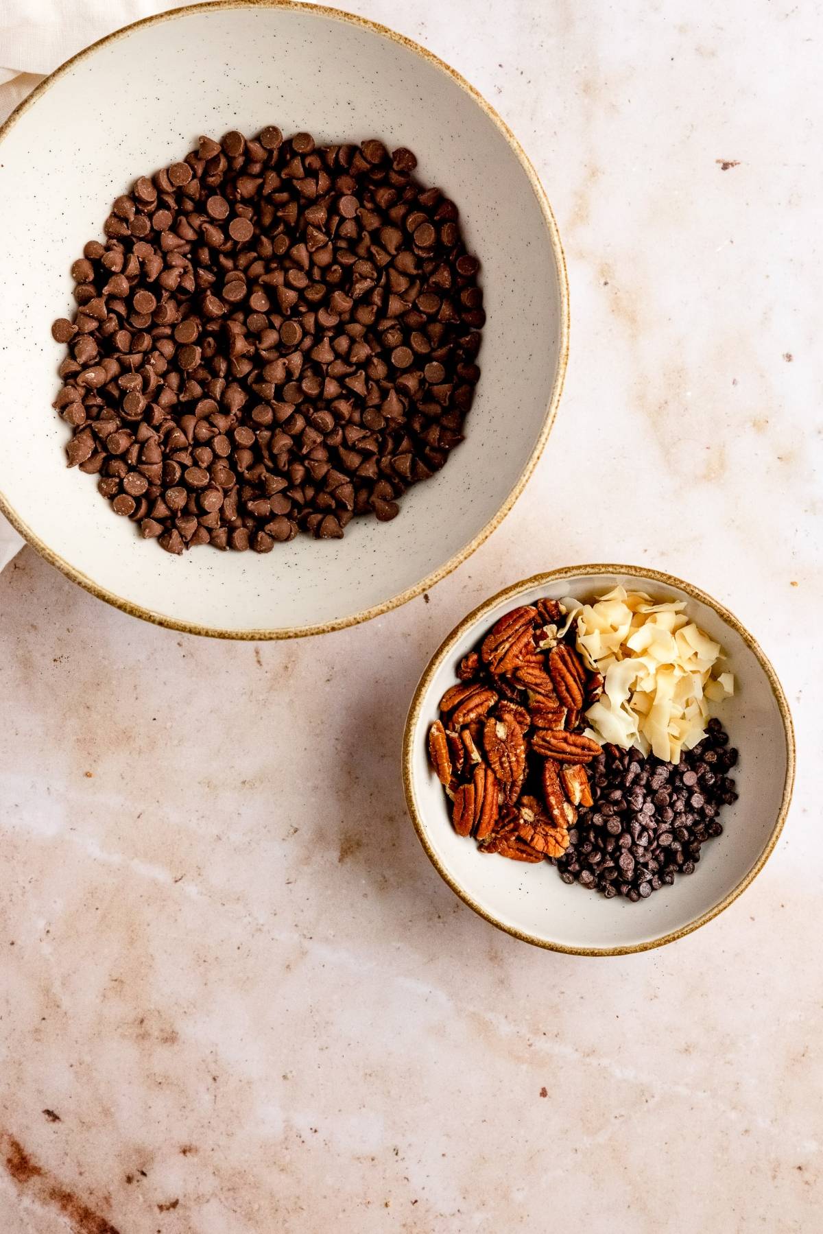 Two bowls: one with chocolate chips, the other featuring cranberry nut clusters with pecans, almond slices, and mini chocolate chips on a marble surface.