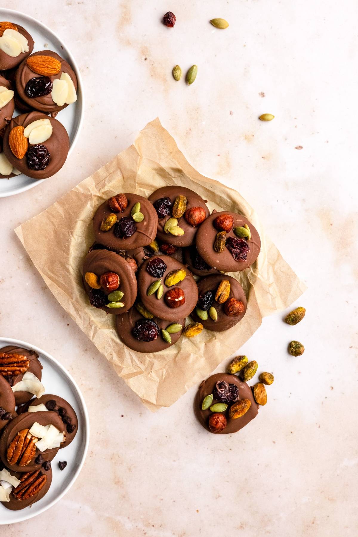 Chocolate discs topped with assorted nuts and dried fruits, these cranberry nut clusters are artfully arranged on parchment and plates on a light surface.