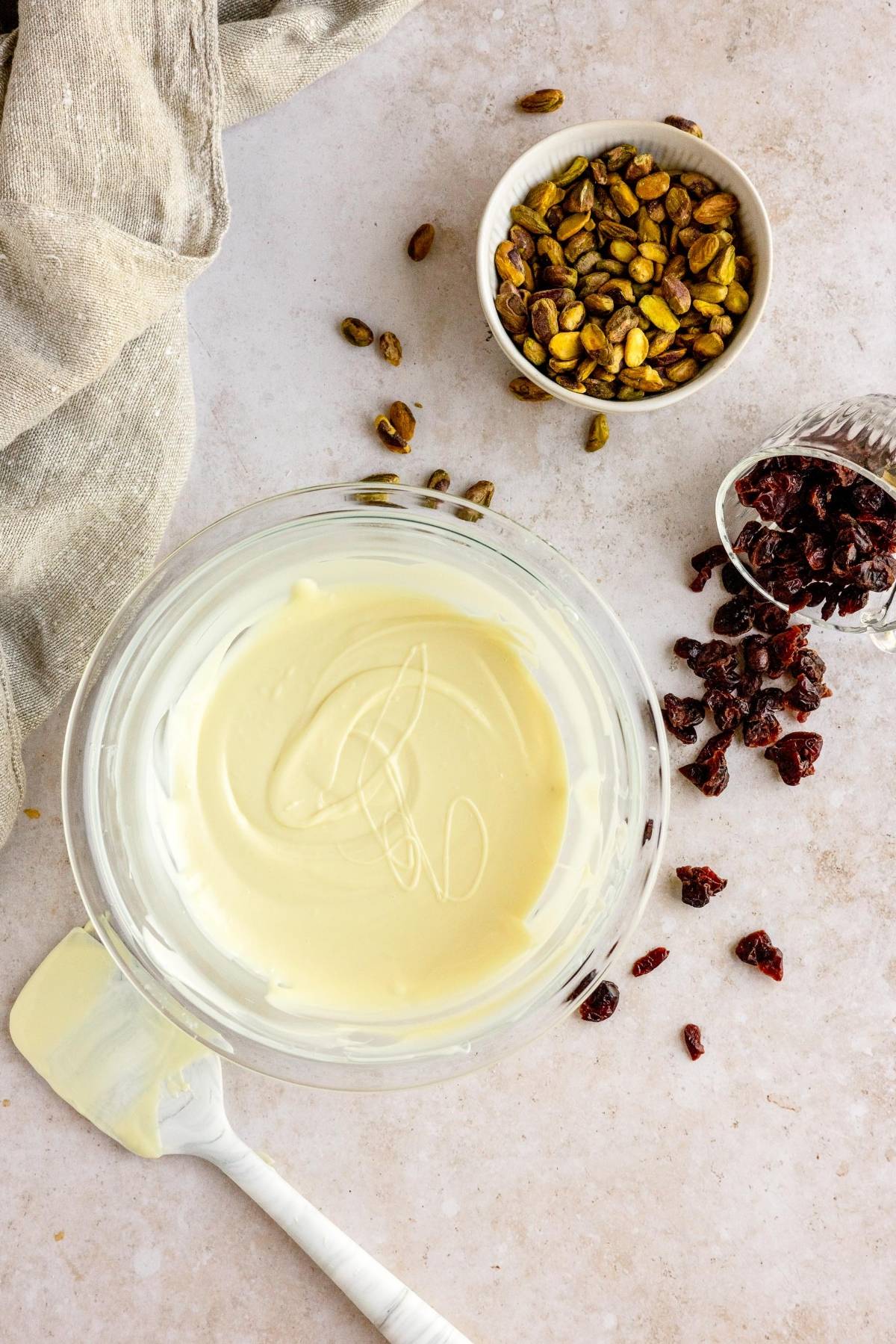 Bowl of melted white chocolate with a spatula, pistachios, and dried cranberries on a countertop—perfect for making Cranberry Pistachio White Chocolate Bark, a treat like almond bark.