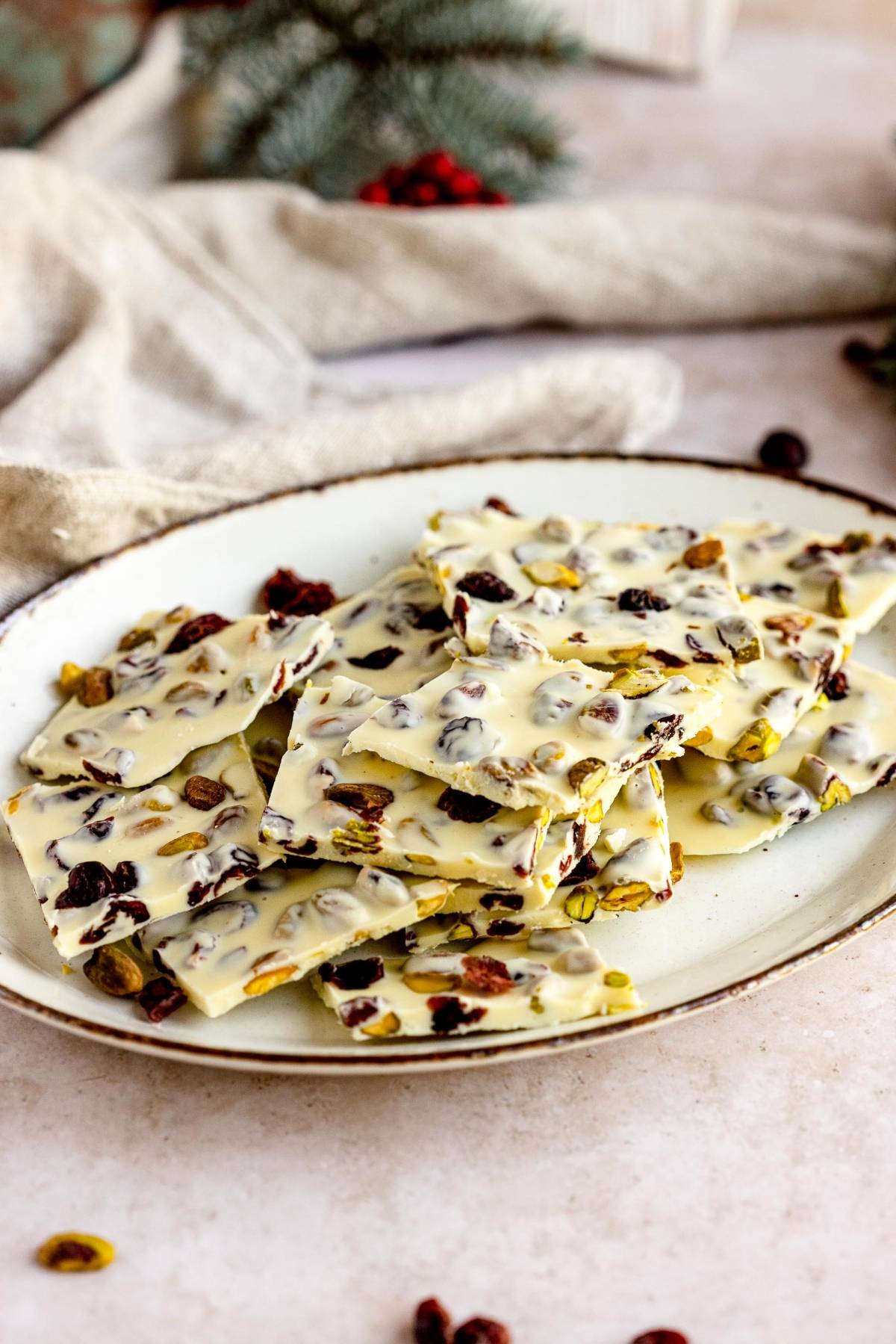 White chocolate bark with nuts and dried fruit, like almond bark, arranged on a white plate with a neutral background. Cranberry and pistachio pieces add festive color and flavor.
