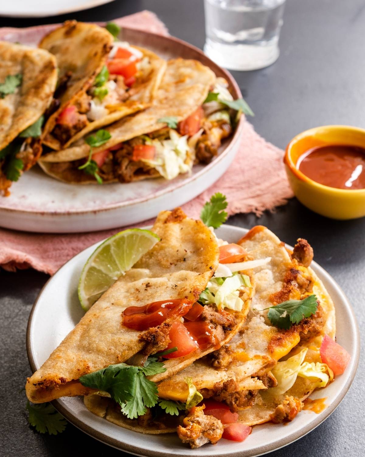 Two plates of crispy tacos filled with meat, lettuce, tomato, cilantro, and cheese, with lime and hot sauce on the side—perfect for a quick and easy dinner or anyone curious about how to make a crispy taco at home.