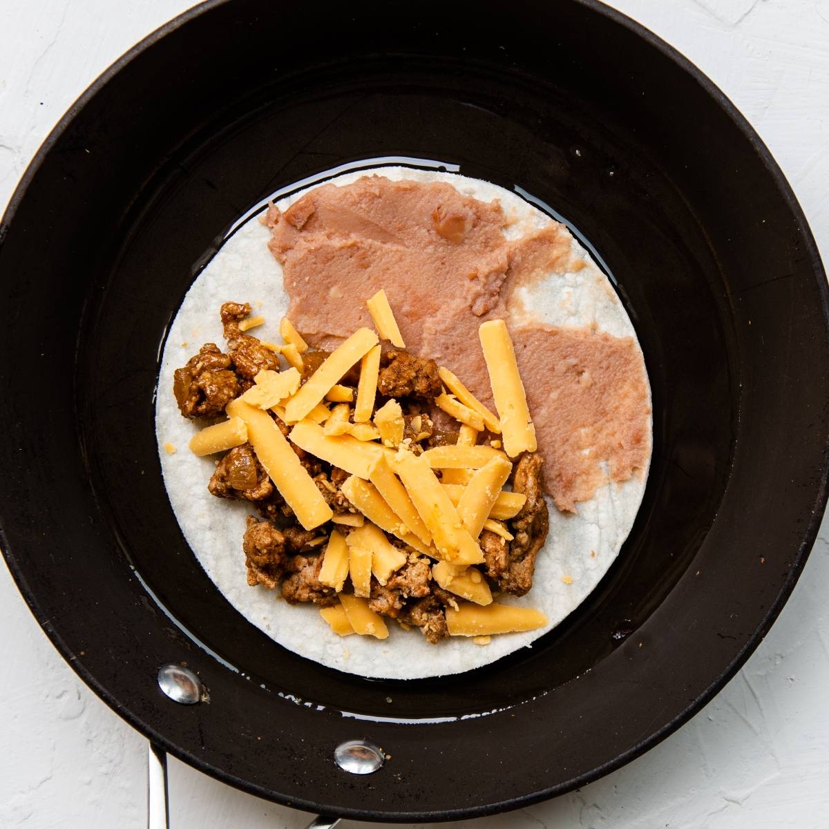 A quick and easy dinner: tortilla in a skillet topped with refried beans, ground turkey, and shredded cheddar cheese—your new favorite turkey taco recipe.