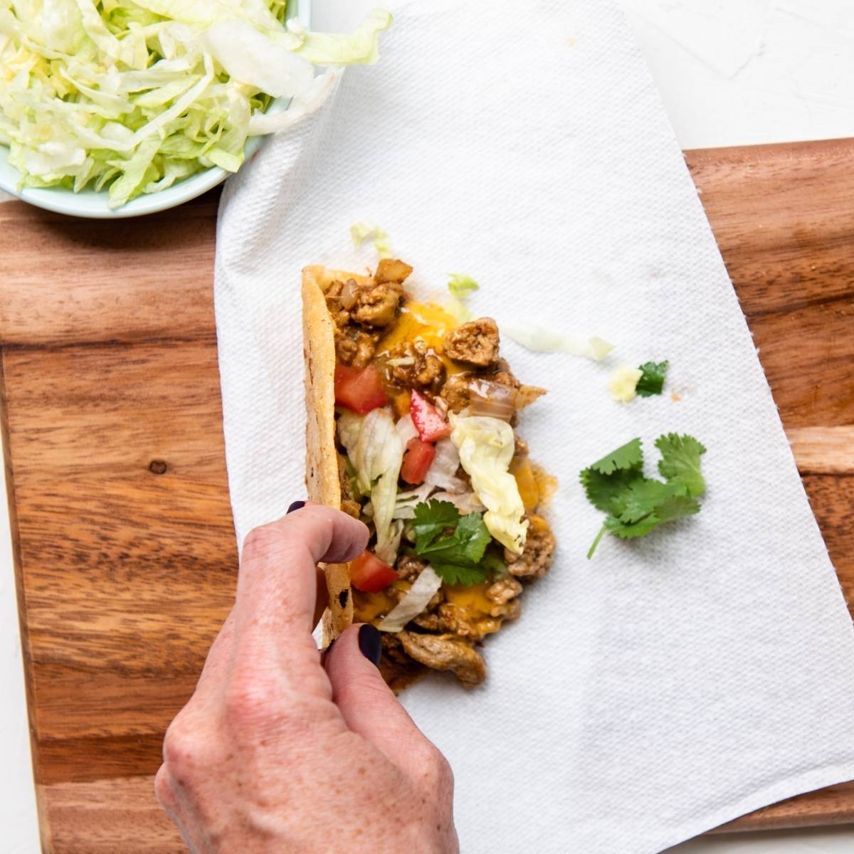 Hand holding a crispy turkey taco filled with meat, lettuce, tomatoes, and cheese on a paper towel, next to a bowl of shredded lettuce—a quick and easy dinner idea.