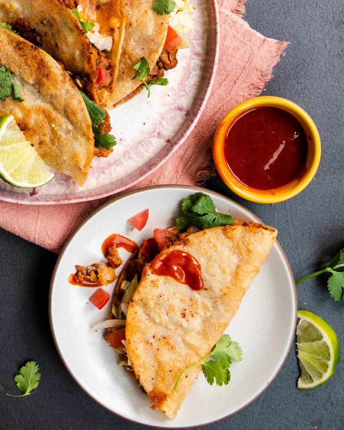 Two plates with crispy turkey tacos, fresh cilantro, lime wedges, and a small bowl of red sauce on the side—perfect for anyone looking for a delicious recipe with ground turkey.