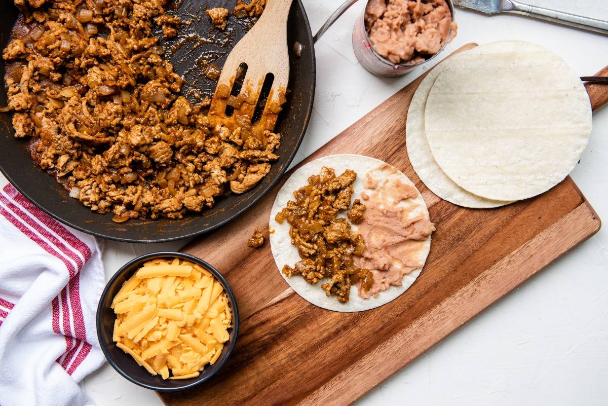 A skillet with taco meat, tortillas, refried beans, and shredded cheese on a wooden board—perfect for a quick and easy dinner or for those learning how to make a crispy taco at home.
