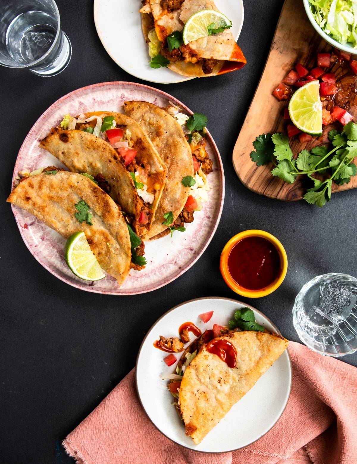 Three crispy tacos with lime and cilantro on plates, served with salsa, lettuce, and tomatoes on the side—a quick and easy dinner for taco night. Try this turkey taco recipe if you want to learn how to make a crispy taco at home!.