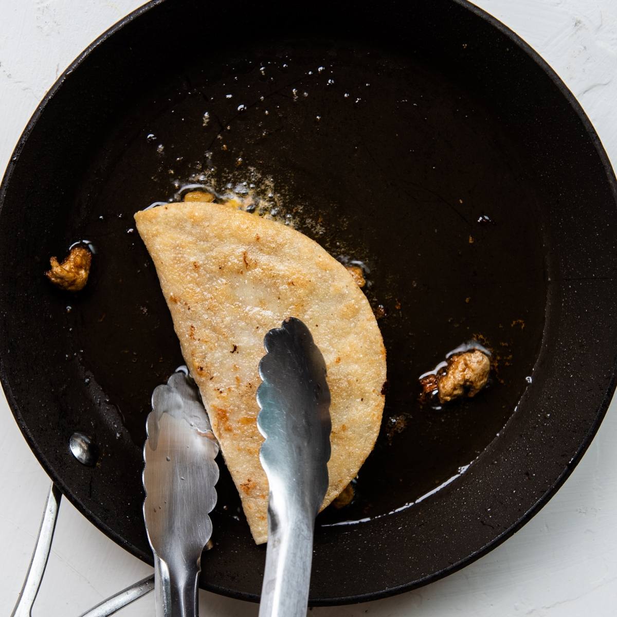A taco is being fried in a skillet with metal tongs pressing it down—a quick and easy dinner and a great recipe with ground turkey for those wanting to learn how to make a crispy taco.