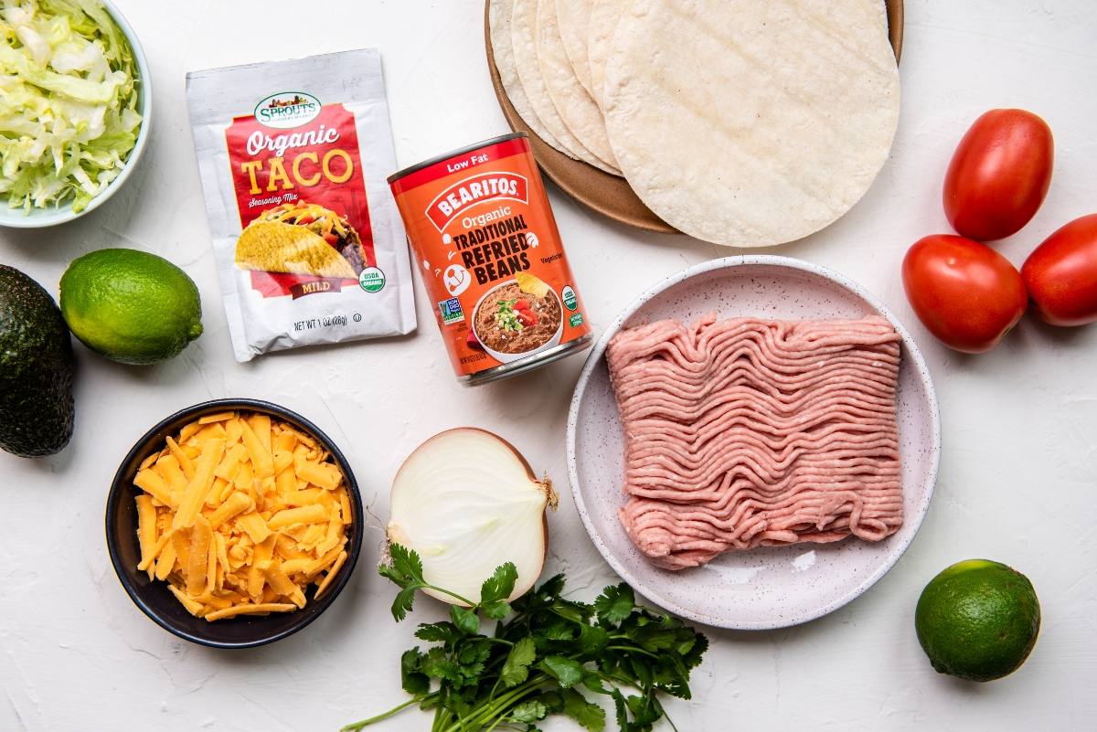Taco ingredients: ground turkey, tortillas, cheese, onion, tomato, taco seasoning, beans, limes, avocado, parsley—a quick and easy dinner recipe with ground turkey for flavorful turkey tacos.