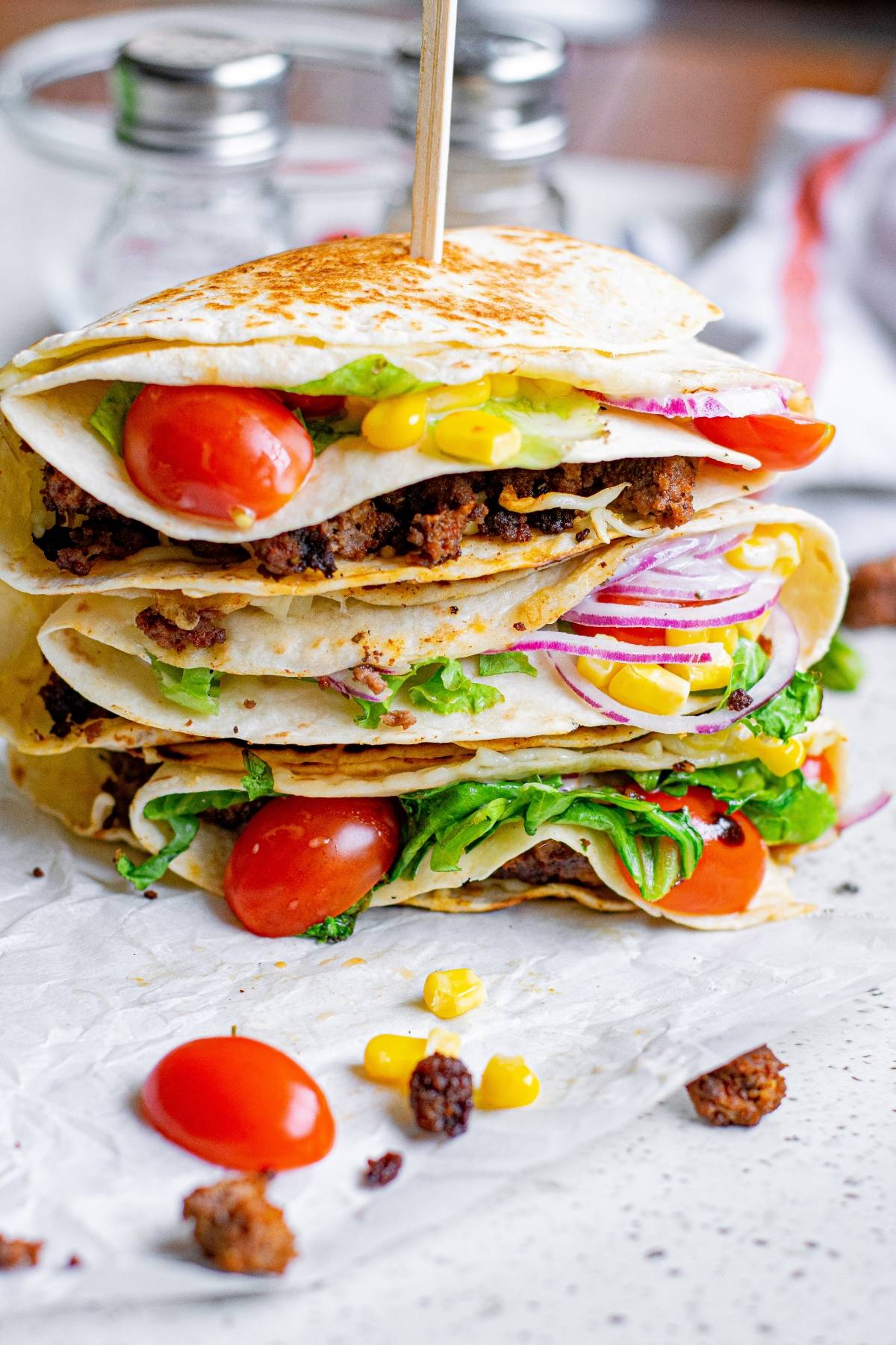 Stacked quesadillas filled with ground meat, lettuce, tomato, corn, and red onion on parchment paper, made using the folded tortilla hack for a fun twist.