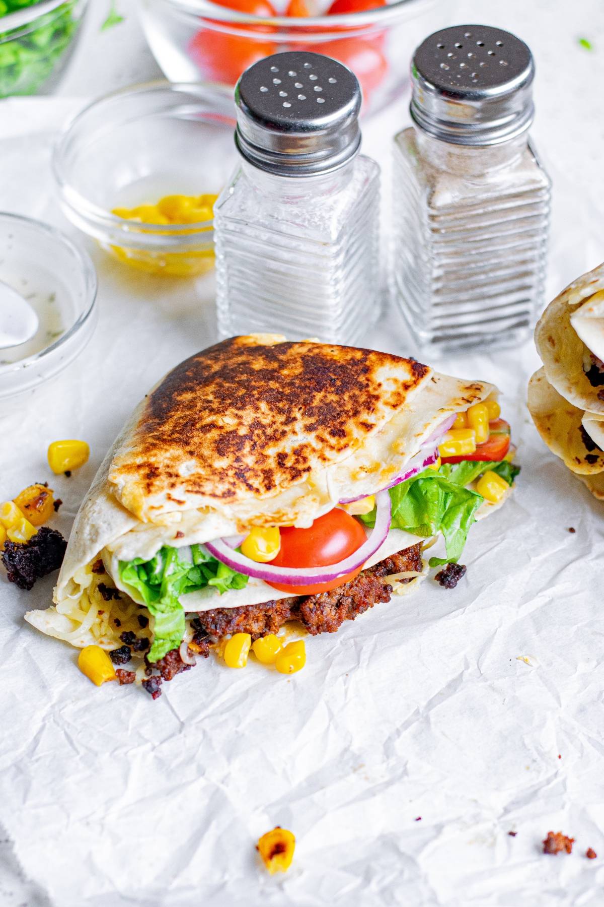 A grilled wrap inspired by the Tik Tok folded tortilla trend, filled with lettuce, tomato, onions, corn, and a crispy patty, served next to salt and pepper shakers.