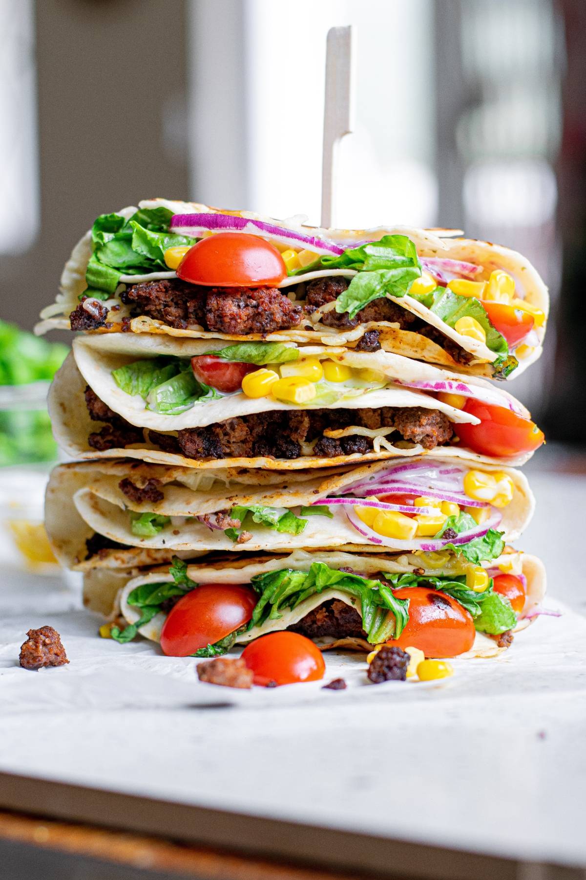 A stack of three stuffed tacos, inspired by the TikTok folded tortilla trend, filled with lettuce, tomatoes, corn, onions, and ground meat on a white surface.