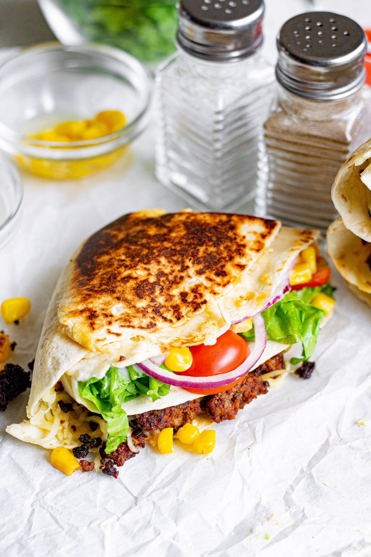 A crispy TikTok folded tortilla wrap with lettuce, tomato, red onion, corn, and meat sits next to salt and pepper shakers.