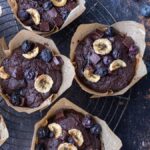 Five Double Chocolate Blueberry Banana Muffins with banana chips and blueberries on a wire rack, surrounded by scattered blueberries and cocoa.