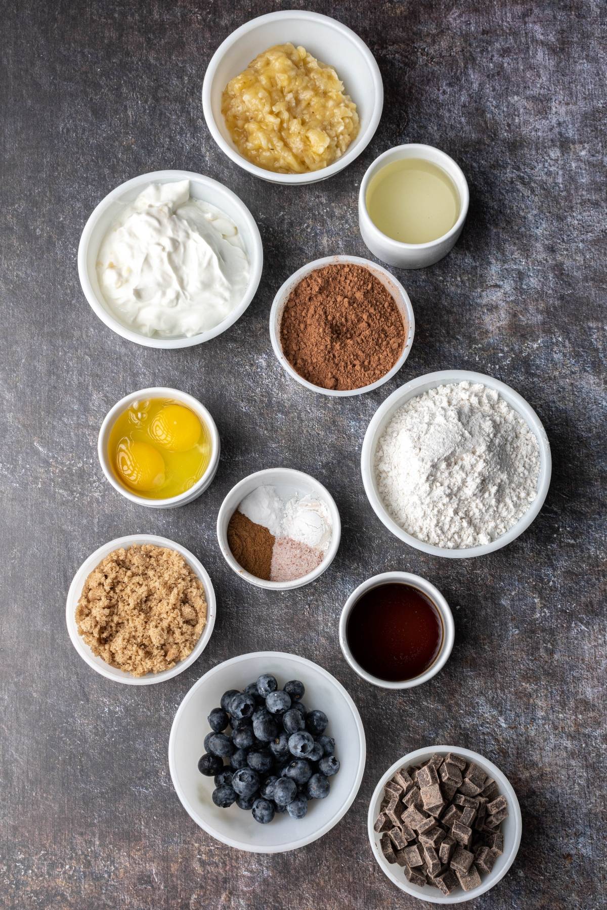 Overhead view of baking ingredients in bowls: eggs, flour, blueberries, bananas, chocolate, cocoa, and more.