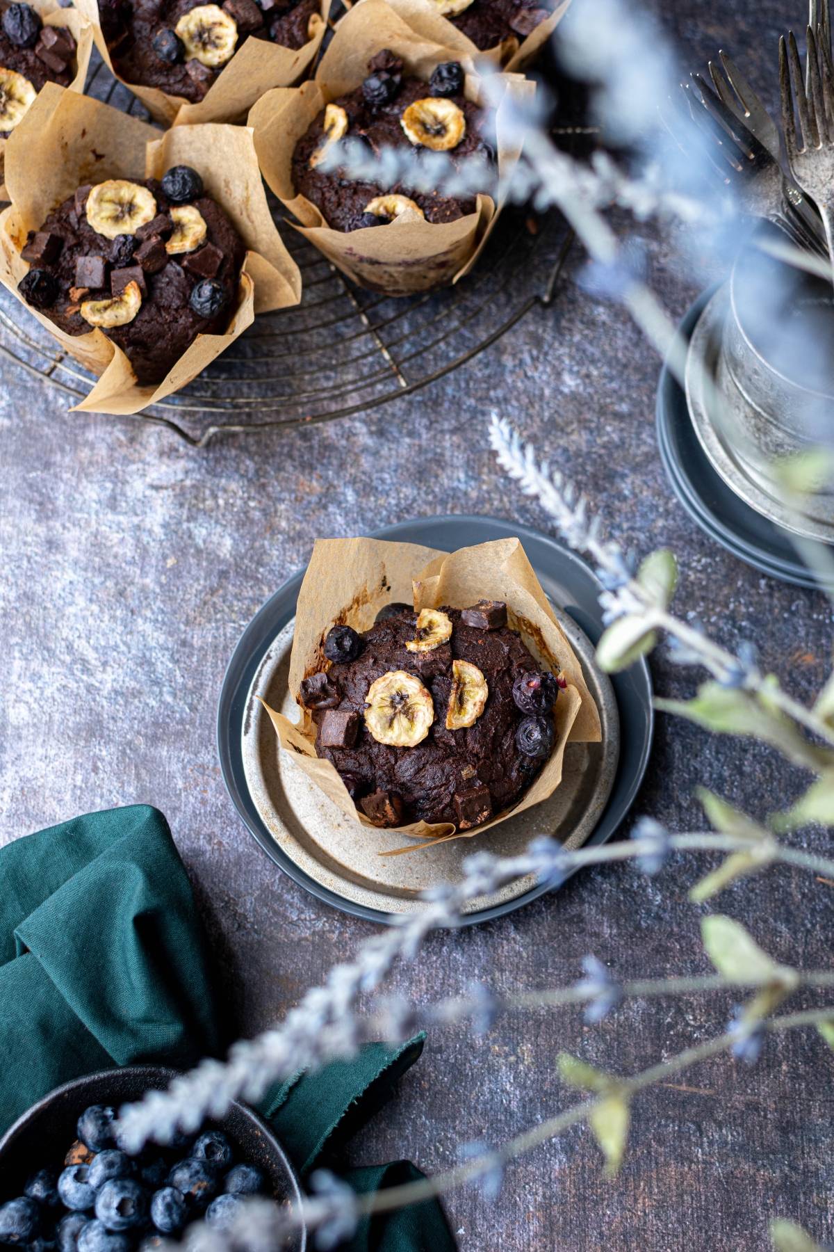 Chocolate muffins with banana chips and blueberries on a cooling rack and plate, with a green napkin nearby.