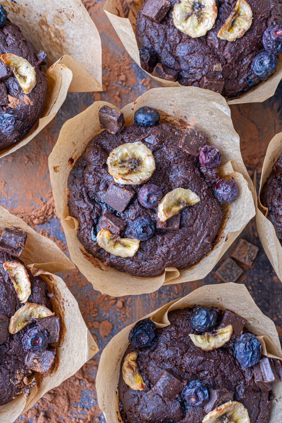 Chocolate muffins topped with banana chips, blueberries, and chocolate chunks in brown parchment paper liners.