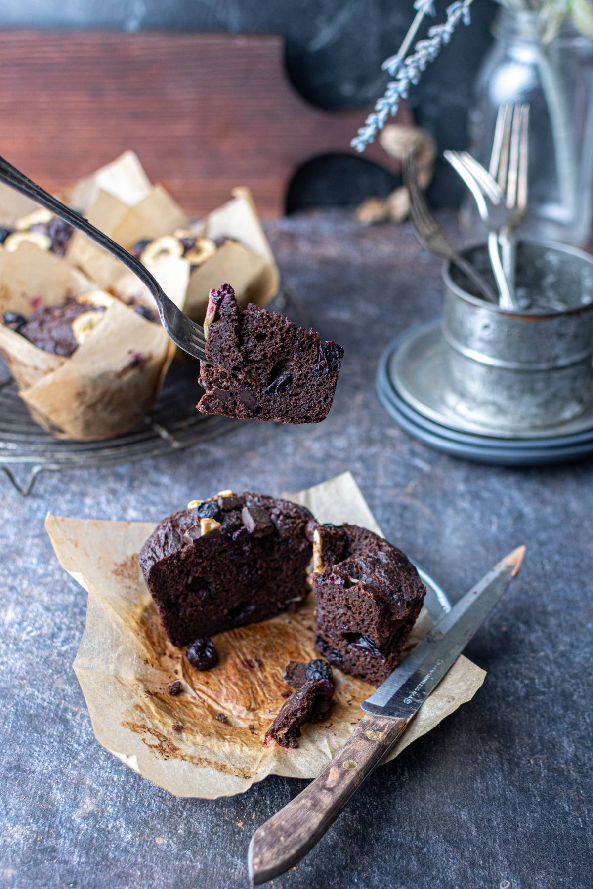 A fork holds a bite of chocolate muffin above a cut muffin on parchment paper with a knife nearby.