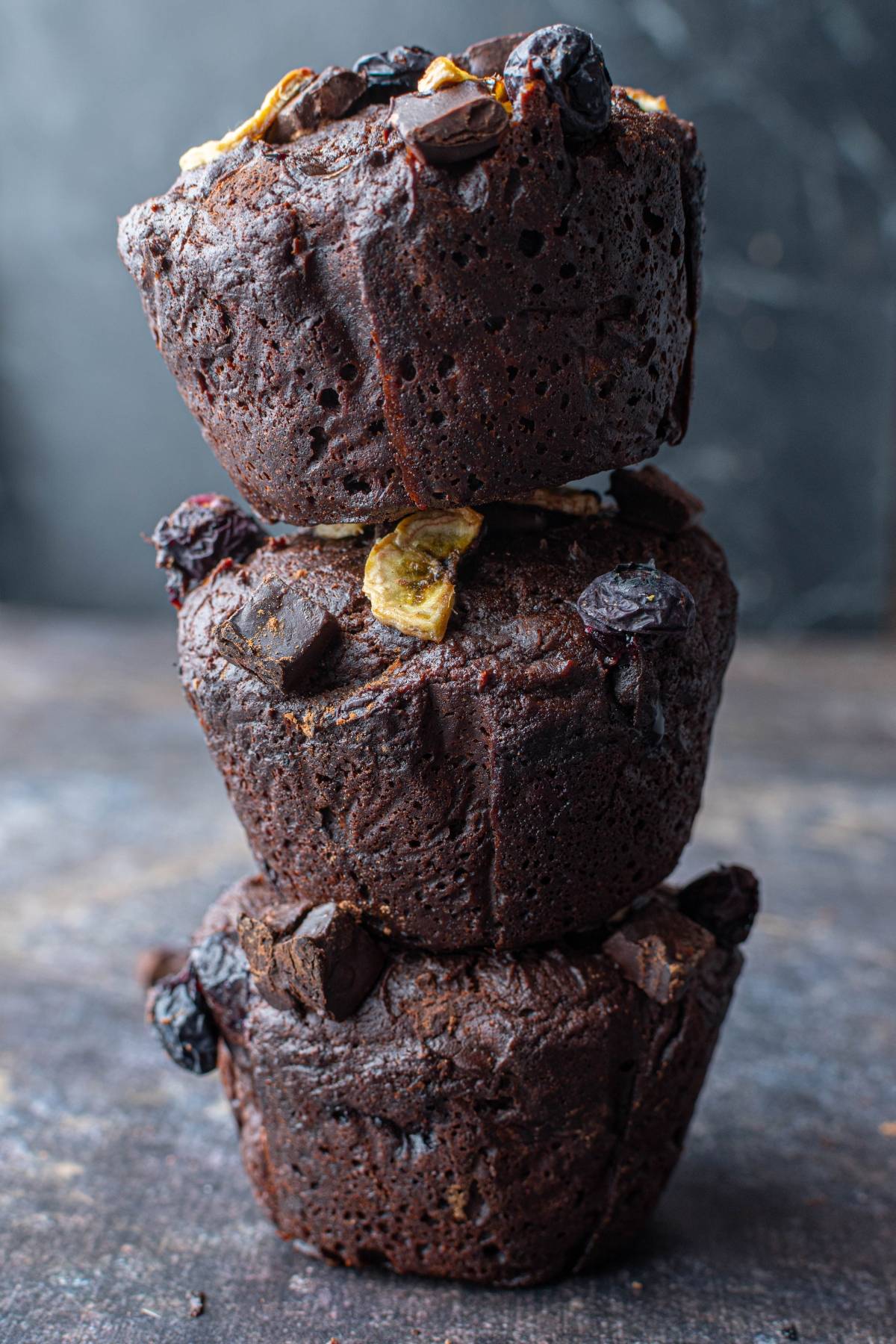 Three stacked chocolate muffins with chocolate chunks and dried fruit on top, set against a dark background.