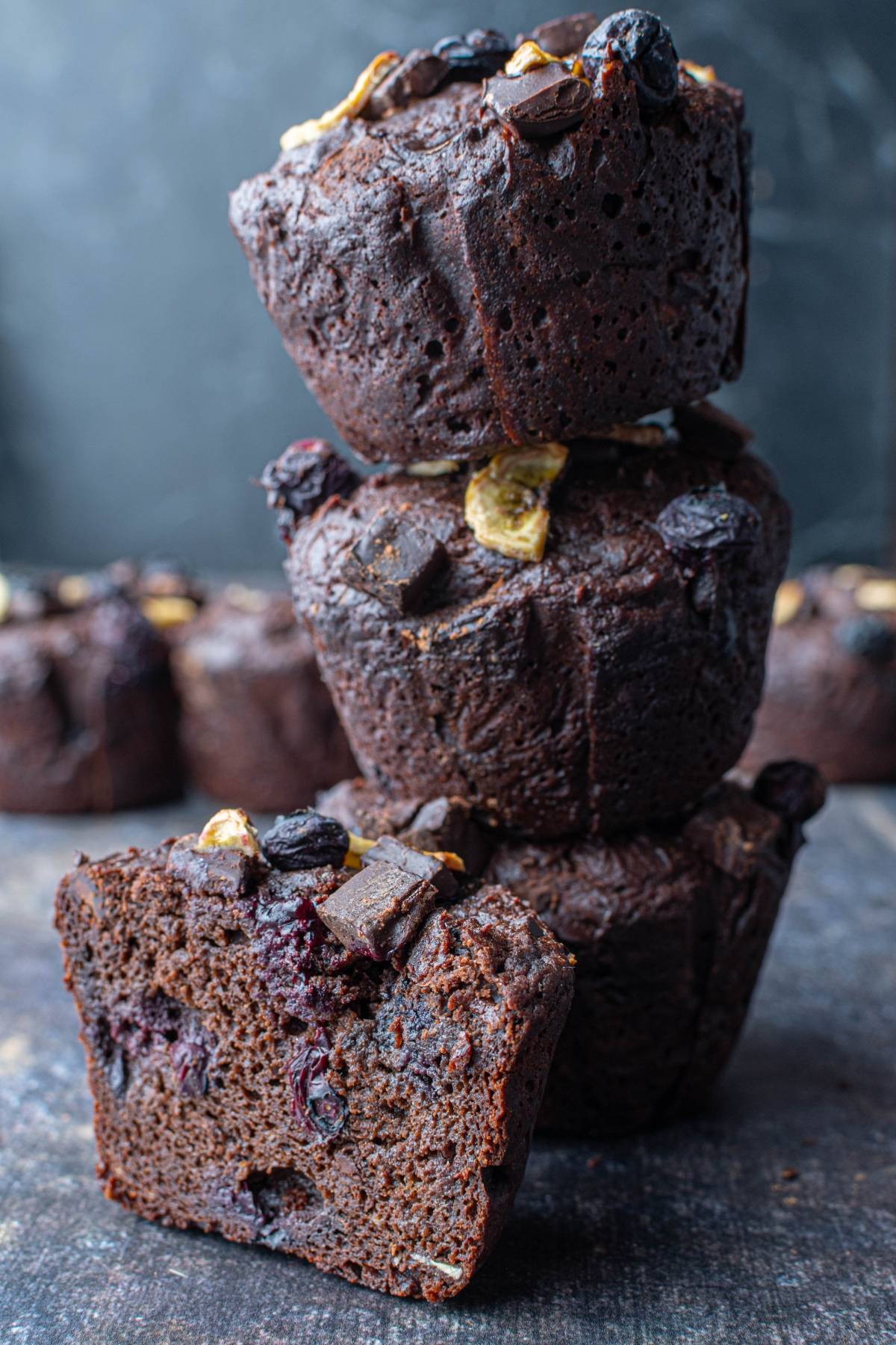 Three stacked chocolate muffins with fruit and chocolate chunks, one muffin cut in half, on a dark surface.