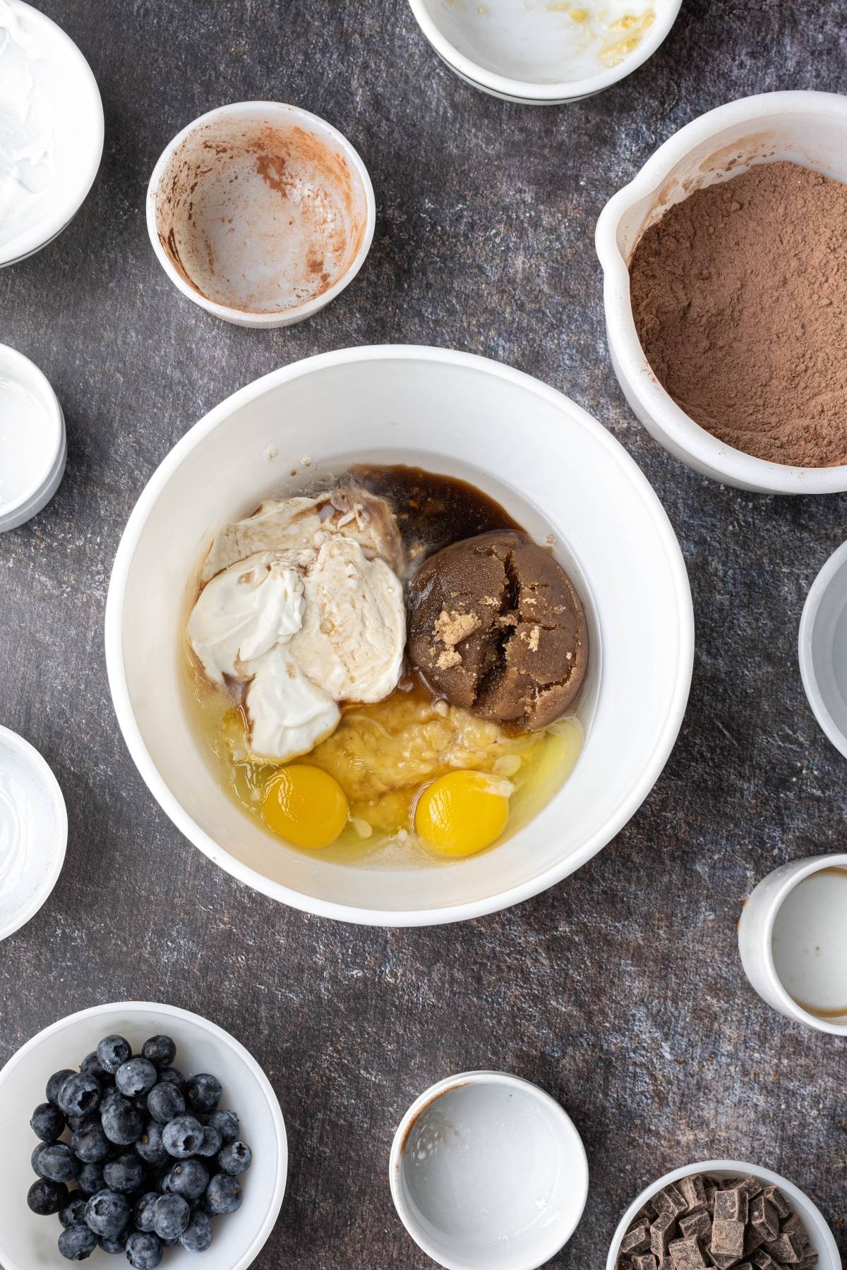 A mixing bowl with eggs, brown sugar, yogurt, and banana, surrounded by bowls of cocoa, blueberries, and chocolate.