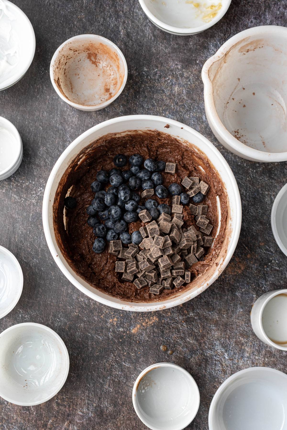A mixing bowl with chocolate batter, blueberries, and chocolate chunks, surrounded by empty bowls.