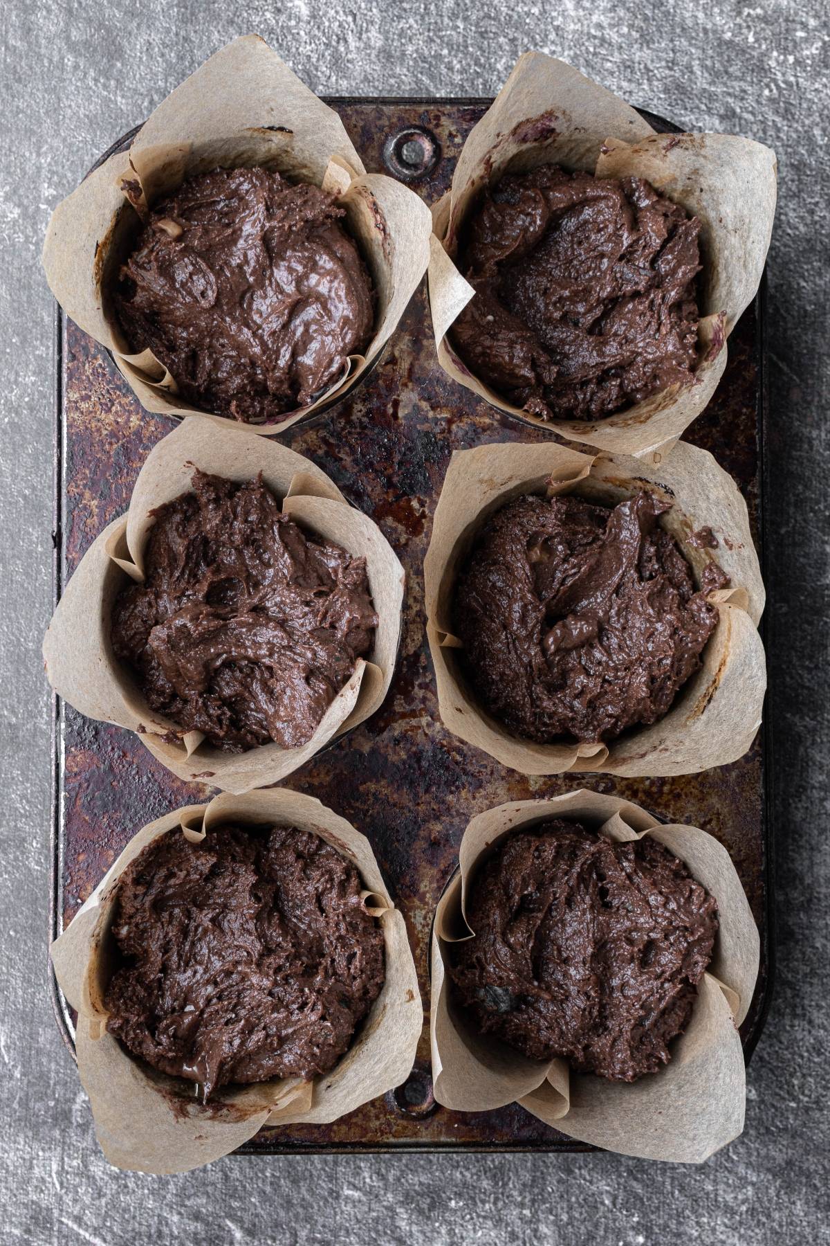 Six unbaked chocolate muffin batters in parchment liners on a rustic muffin tray, ready for baking.