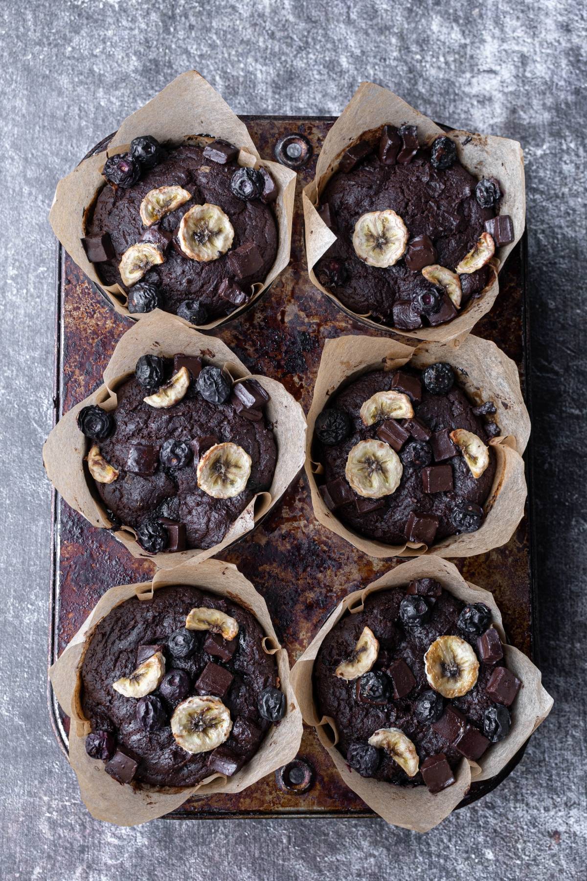 Six chocolate muffins with banana slices and blueberries in parchment paper on a rustic metal tray.