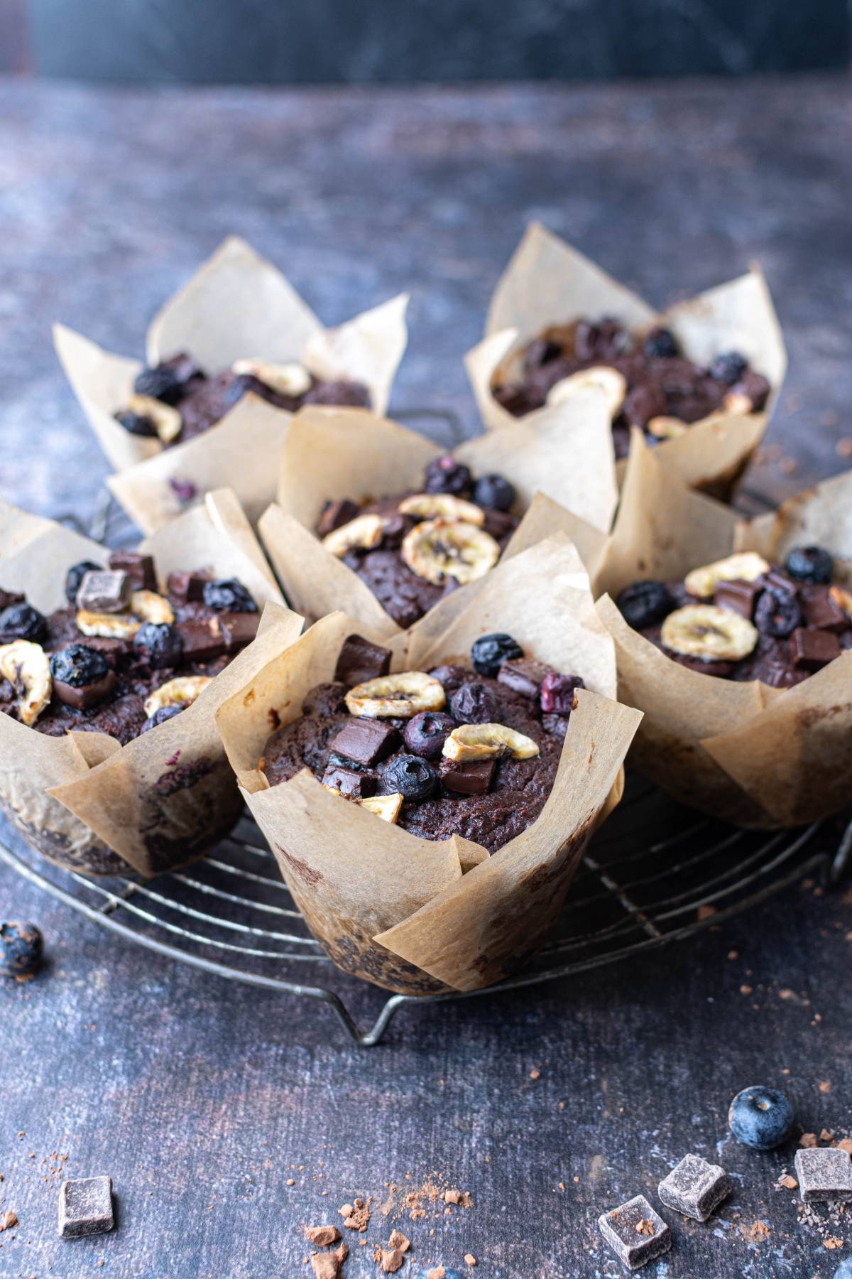 Six chocolate muffins with blueberries and banana slices on top, sitting on a wire rack with scattered toppings.