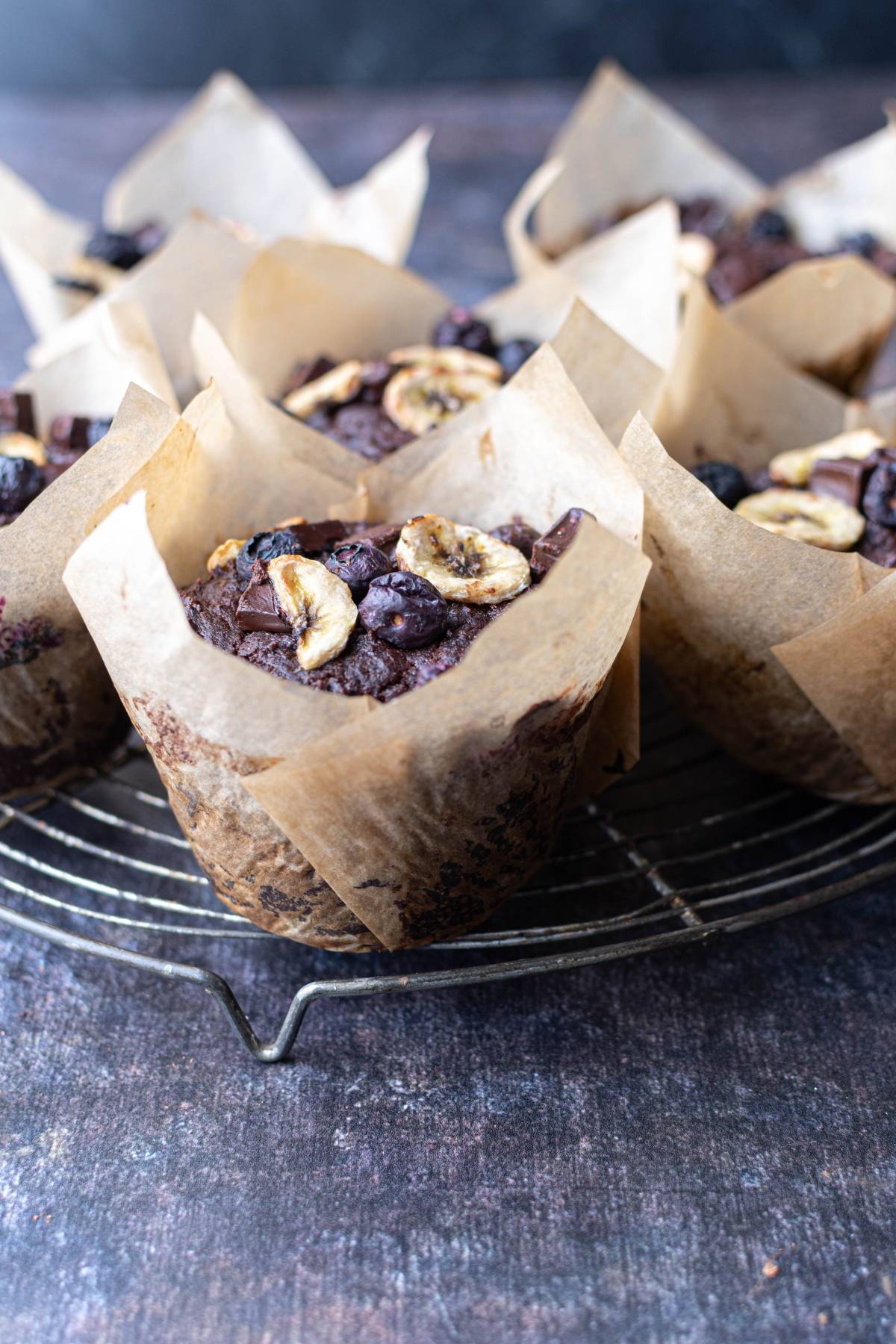 Chocolate muffins with dried banana slices and chocolate chunks in parchment wrappers on a cooling rack.