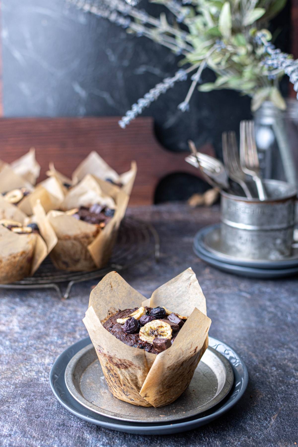 A chocolate and banana muffin in parchment on a plate, with more muffins and rustic decor in the background.