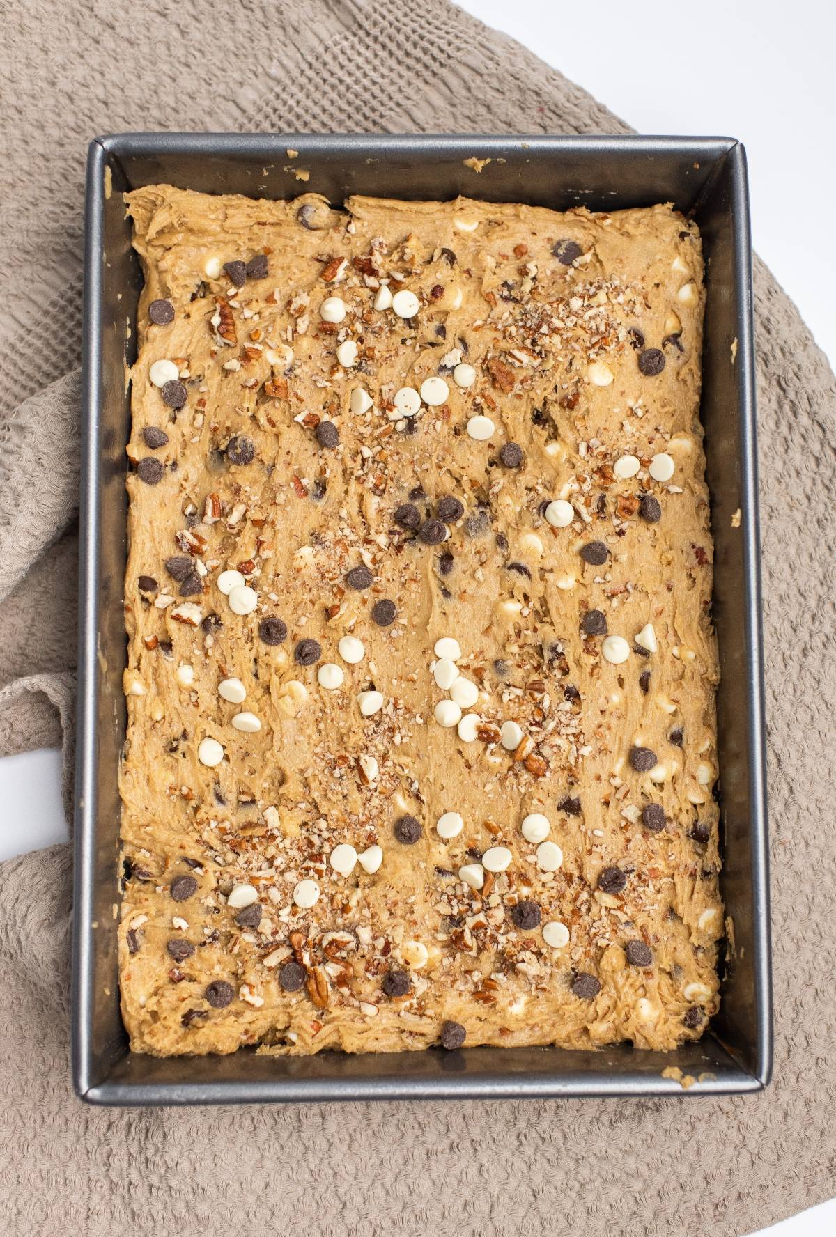 Rectangular pan of unbaked Double Chocolate Pecan Blondies batter topped with chocolate chips and nuts on a textured cloth.