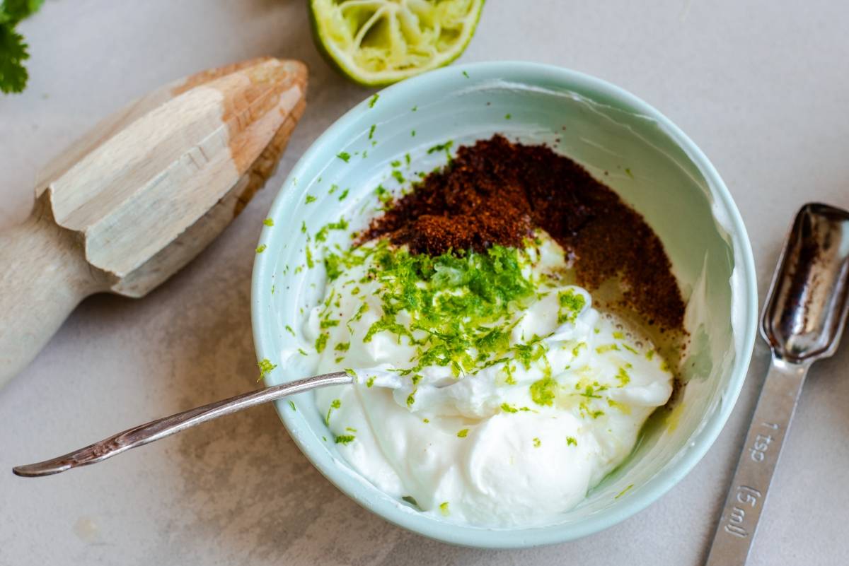A bowl of yogurt with spices and lime zest sits beside a spoon, juicer, and lime half—the perfect creamy topping for an Elote Corn Kale Salad or any Mexican inspired salad.